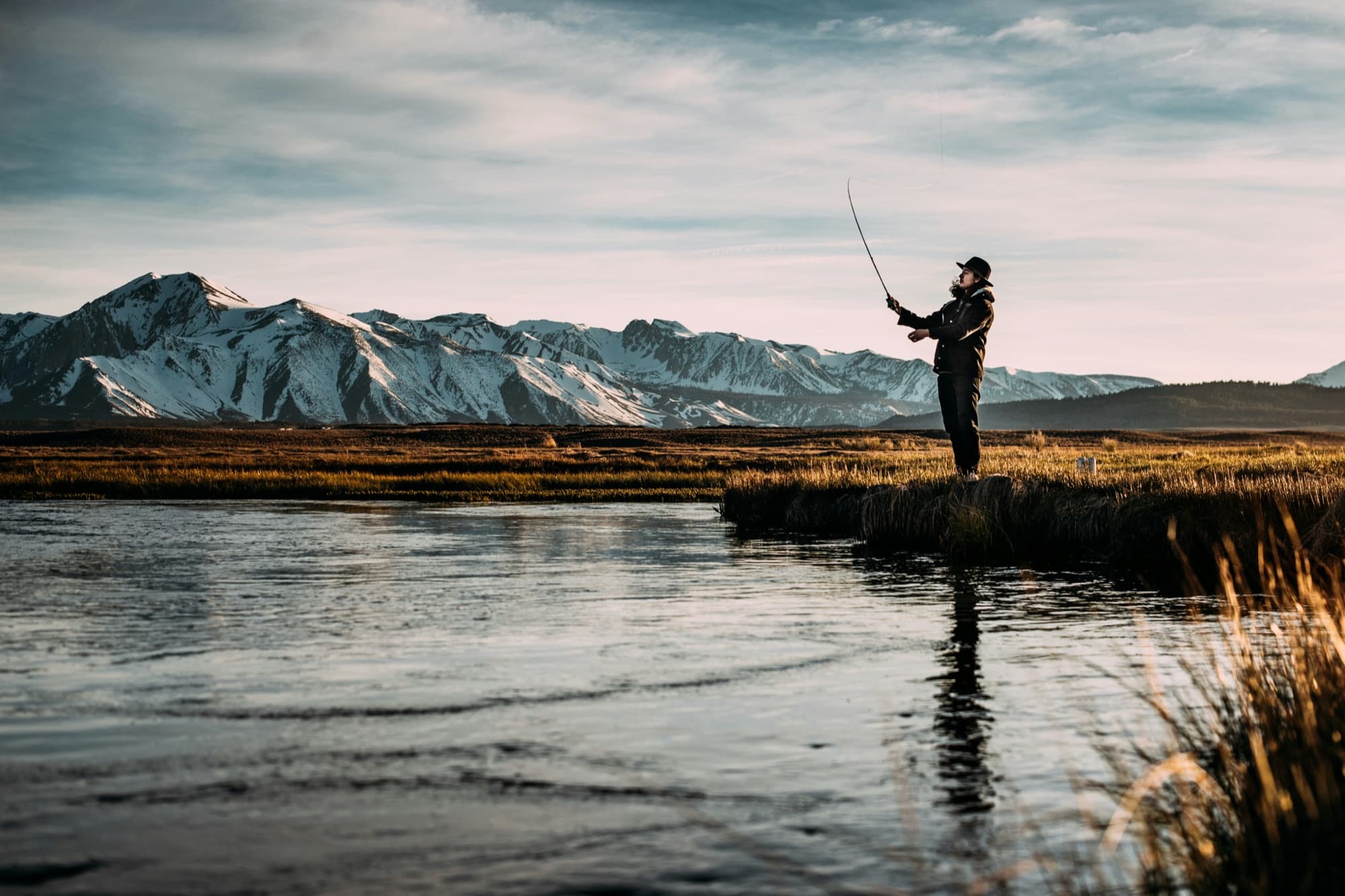 A fly fisherman casting a line into a calm river at sunrise with snow-capped mountains behind