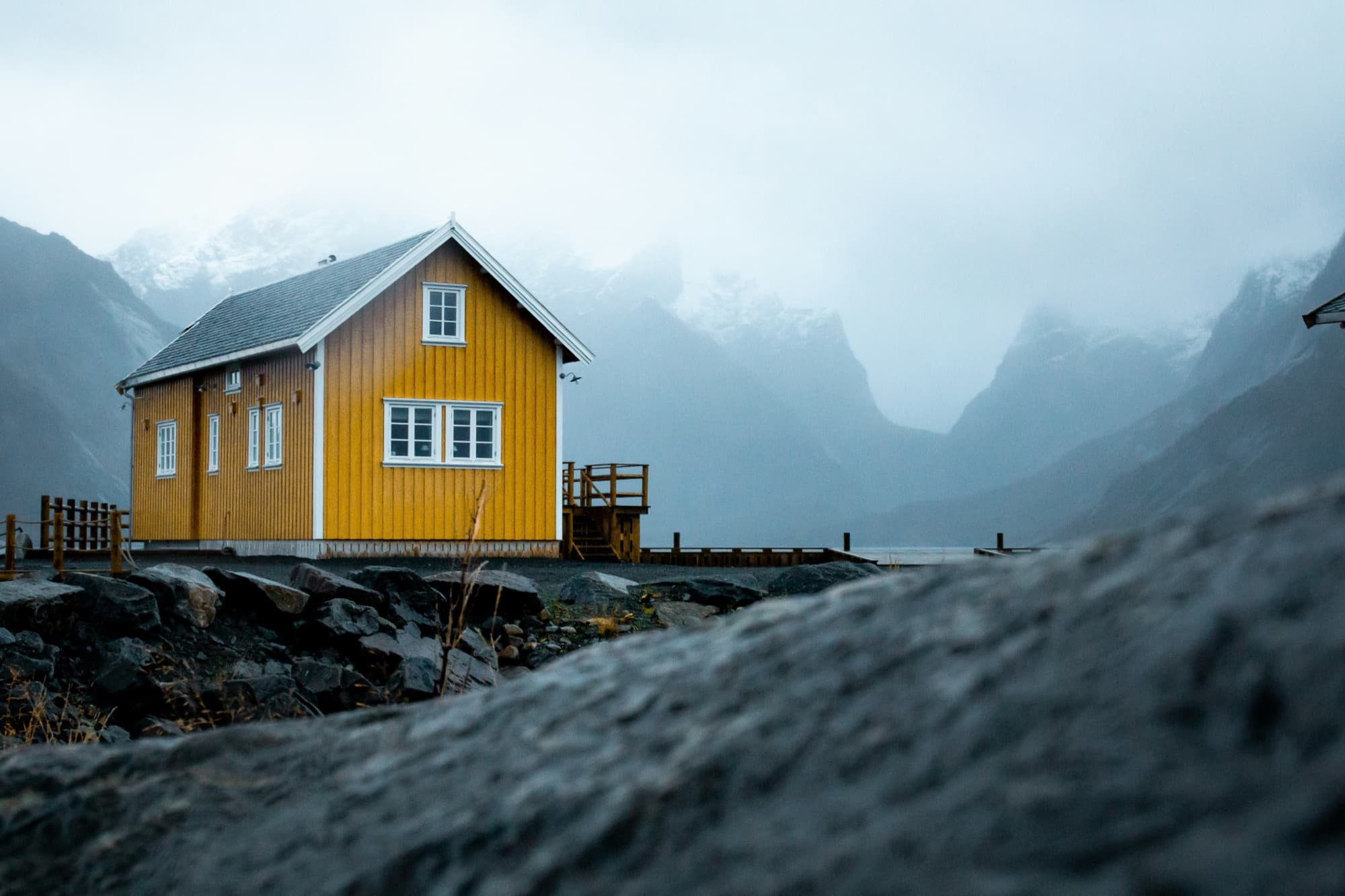 A small yellow wooden cabin standing on rocky ground in front of misty, snow-dusted mountains