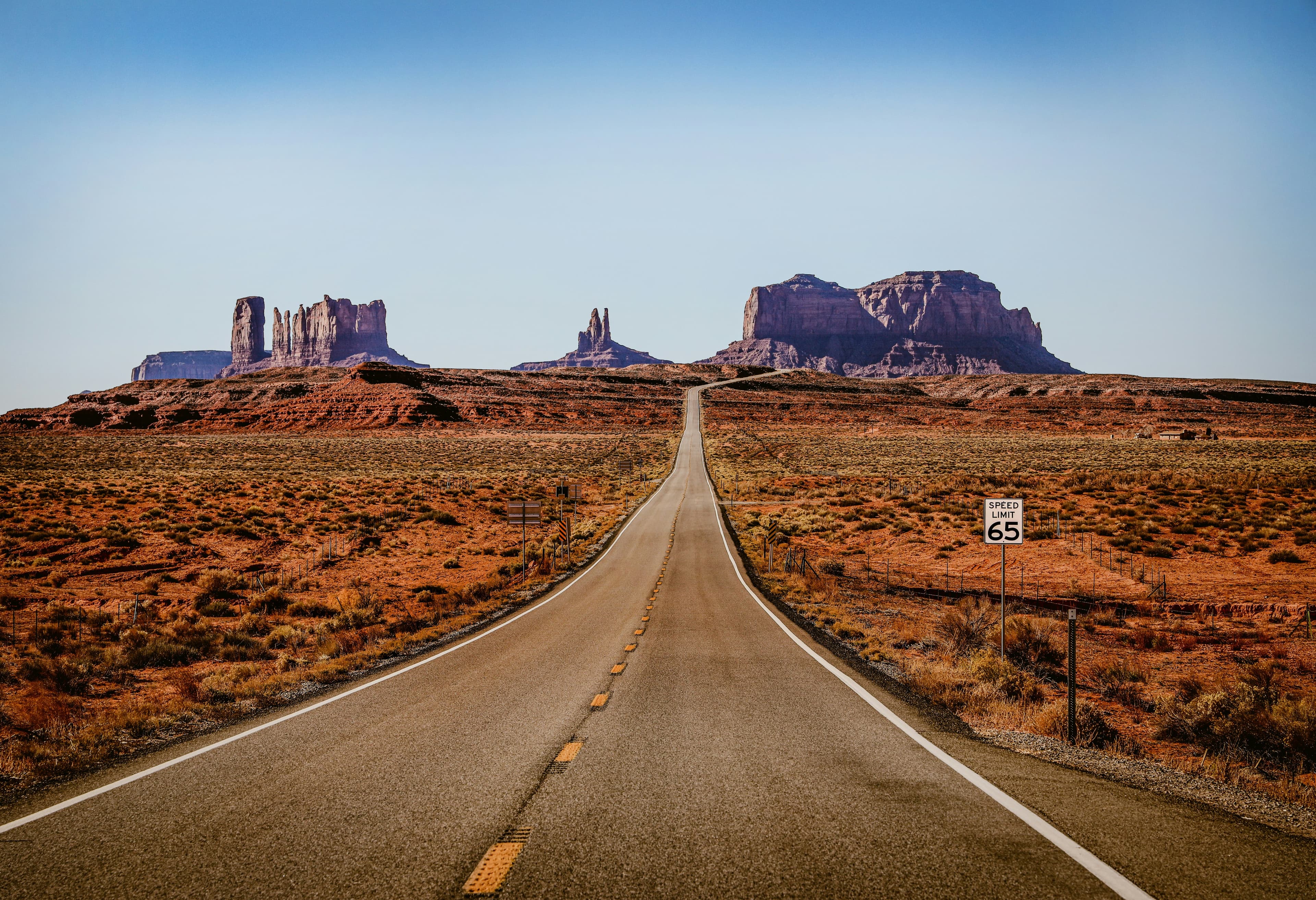 An empty desert highway stretching straight toward the red buttes of Monument Valley under a soft blue sky
